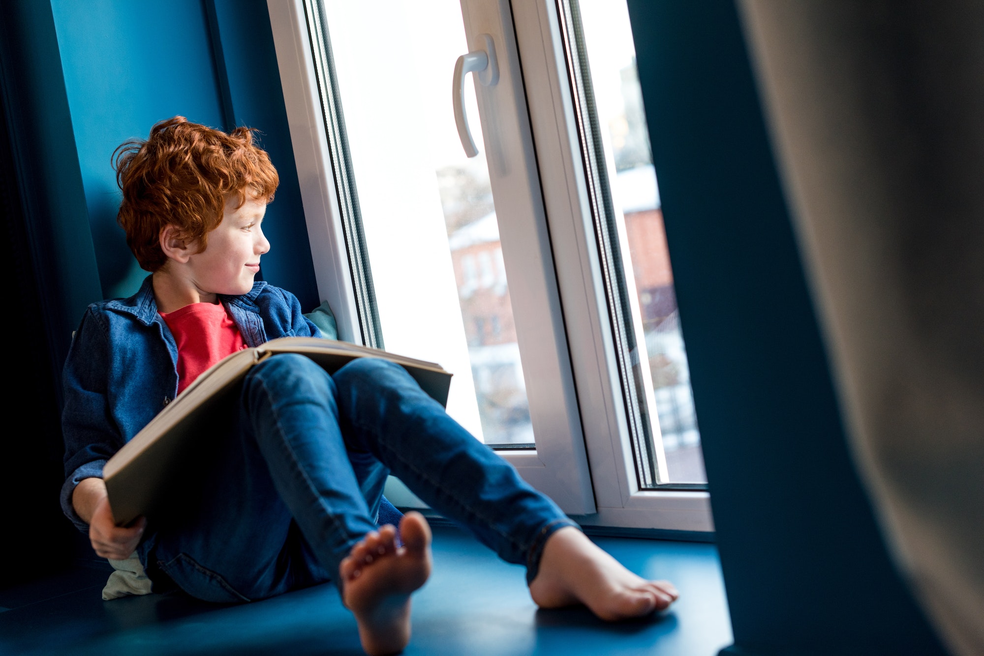 cute barefoot boy with book sitting on windowsill